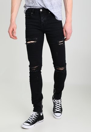 Person trägt schwarze zerrissene Skinny-Jeans und schwarze High-Top-Sneaker mit weißen Schnürsenkeln vor einfarbigem Hintergrund.