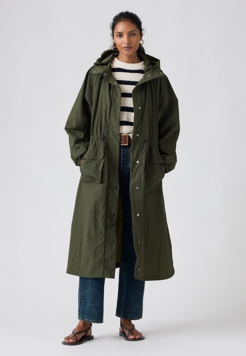 Imperméable long vert olive avec une taille à cordon de serrage, des pressions et de grandes poches avant, porté sur un pull rayé et un jean à jambes larges.