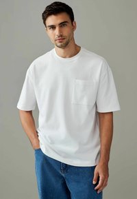 Hvid t-shirt med korte ærmer, rund halsudsk�æring og en lomme på brystet. Stoffet ser blødt og glat ud, parret med blå denim shorts.