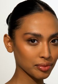 Nærbilde av en person med middels hudtone, jevn tekstur, mørkt hår i en knute, brune øyne og en nøytral leppfarge mot en hvit bakgrunn.
