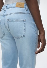 Ανοιχτό μπλε τζιν παντελόνι με πίσω τσέπες και καφέ δερμάτινο ταμπελάκι με την επιγραφή "ONLY Blue Denim", φορεμένο από άτομο με σκουρόχρωμο δέρμα και γκρι μπλούζα.