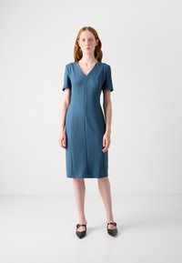Teal short-sleeve fitted dress with a V-neckline, knee-length hem, and vertical seams, paired with black pointed flats.