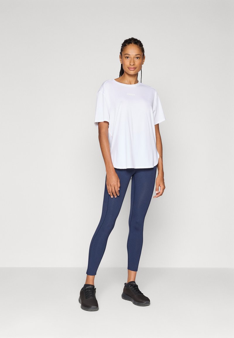 Frau mit geflochtenem Haar, die ein weißes Sportshirt, navyfarbene Leggings und schwarze Turnschuhe trägt, posiert mit einem selbstbewussten Lächeln vor einem neutralen Hintergrund.