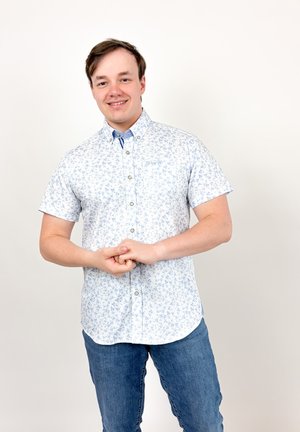 Jeune homme souriant, portant une chemise boutonnée à manches courtes bleu clair à motif fleuri et un jean bleu, debout devant un fond blanc uni.