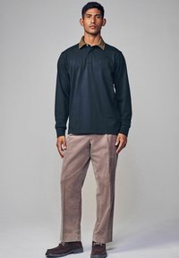 Polo de culoare verde închis cu mânecă lungă, cu un guler din catifea bej, asortat cu pantaloni din catifea bej deschis și pantofi din suede maro închis.