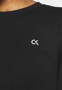 Chemise noire en coton à manches longues avec un col rond et un petit logo CK blanc sur le côté gauche de la poitrine. Texture douce et lisse.