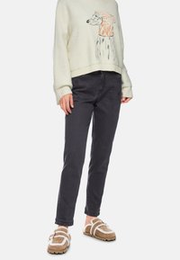 Cremefarbener Pullover mit Bären-Graphic; dunkelgraue Jeans; braune, flauschige Slipper mit weißen Akzenten und Schnallen-Detail.