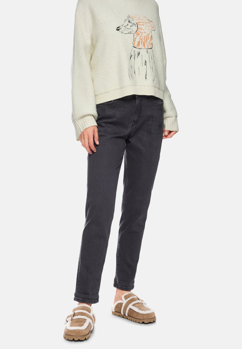 Cremefarbener Pullover mit Bären-Graphic; dunkelgraue Jeans; braune, flauschige Slipper mit weißen Akzenten und Schnallen-Detail.