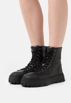 Bottes de combat montantes en cuir noir avec semelles épaisses à relief portées sur des jambes nues sur un fond blanc.