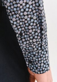 Czarna rękawka koszuli z kwiatowym wzorem, na którym znajdują się małe niebieskie i różowe kwiaty. Na mankiecie znajduje się detal w postaci guzika oraz fakturowana tkanina.