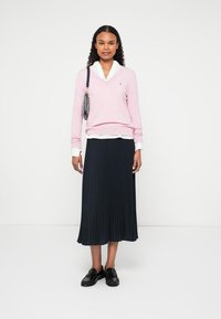 Maglione rosa a scollo a V sopra una camicia bianca con colletto, abbinato a una gonna plettata blu navy e scarpe stringate nere. Borsa a tracolla con nappina.