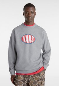Felpa grigia con un grande logo "VANS" rosso e blu. Presenta un colletto a girocollo e maniche lunghe, sovrapposta a una maglietta rossa e pantaloni con motivo mimetico.