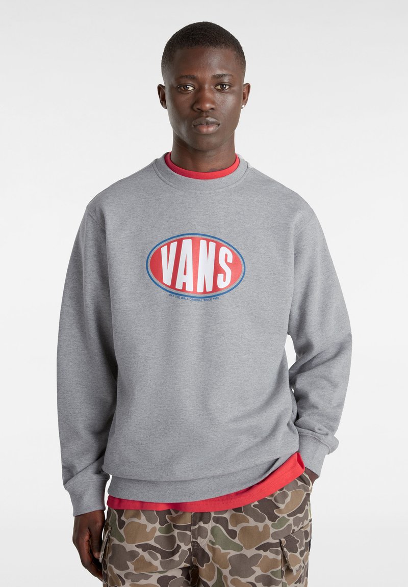 Felpa grigia con un grande logo "VANS" rosso e blu. Presenta un colletto a girocollo e maniche lunghe, sovrapposta a una maglietta rossa e pantaloni con motivo mimetico.