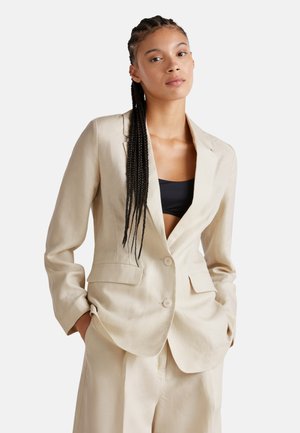 Beige linen blazer with a notched lapel, two front pockets, and two buttons. Worn over a black top, featuring long braids. Smooth texture.