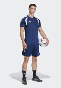 Νεαρός άνδρας σε ναυτική αθλητική ένδυση holding μια λευκή και κόκκινη μπάλα ποδοσφαίρου, φορώντας μαύρα π�απούτσια ποδοσφαίρου και στέκεται σε ανοιχτό φόντο.