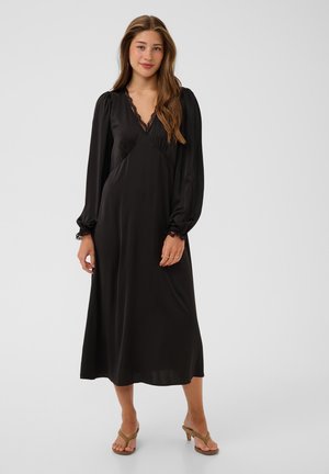 Femme aux longs cheveux bruns portant une longue robe noire avec des détails en dentelle et des sandales à talons beiges, debout devant un fond uni.