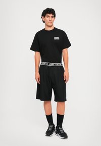 Vyras, dėvintis juodą Versace Couture marškinėlį, šortus su prekiniu juosteliu, juodas kojines ir juodus sportbačius su baltais akcentais ant balto fono.
