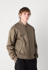 Veste bomber vert olive en tissu lisse et brillant avec une fermeture éclair à l'avant, des poignets côtelés et des poches latérales. T-shirt noir basique en dessous.