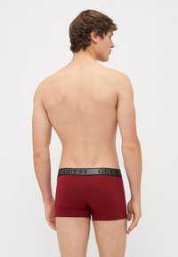 Boxer da uomo bordeaux con elastico nero in vita e logo "GUESS"; tessuto liscio con una vestibilità aderente, mostra una vista posteriore semplice.