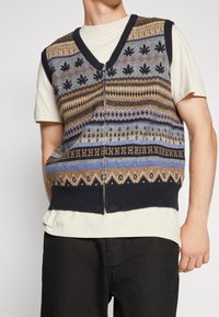 Pletená vesta bez rukávů se zipem, obsahující modré, hnědé a šedé vzory včetně geometrických a listových motivů. Kontrastní tmavě modrý lem.