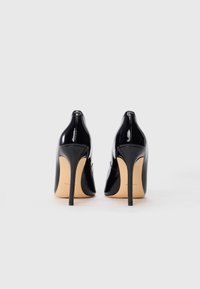 Černé lakované kožené vysoké podpatky s špičatou špičkou a štíhlým stiletto podpatkem, s hladkým povrchem a embosovaným logem na zadní straně.