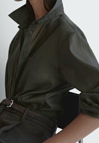 Chemise boutonnée en vert foncé et transparent avec un grand col, une poche poitrine unique et des manches retroussées, associée à une ceinture marron.