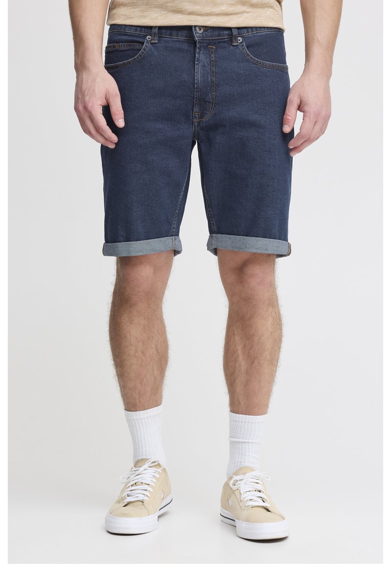 Tummansiniset denimshortsit, joissa on käännetyt lahkeet, viisi taskua ja kontrastiväriset tikkausompeleet. Yhdistetty beigenvärisiin lenkkareihin ja valkoisiin keskipitkiin sukkiin.