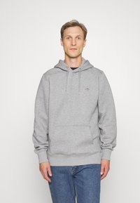 Grå hettegenser med snor og frontlomme, laget av mykt materiale. Kombinert med blå jeans. Enkelt logodesign til stede.