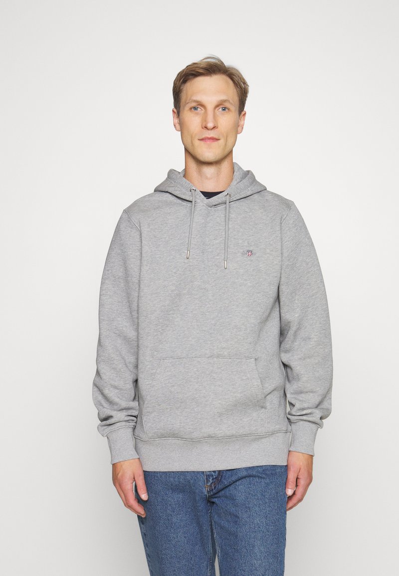 Grå hettegenser med snor og frontlomme, laget av mykt materiale. Kombinert med blå jeans. Enkelt logodesign til stede.