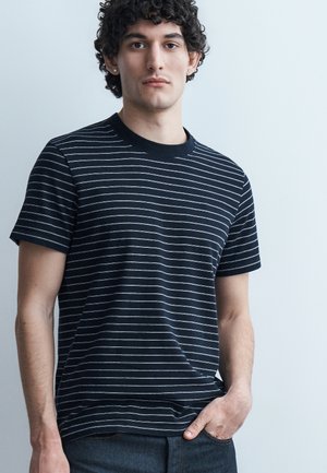 Νεαρός άνδρας με σγουρά μαλλιά που φοράει μαύρο κοντομάνικο πουκάμισο με λεπτές λευκές οριζόντιες ρίγες, στέκεται μπροστά σε ένα απλό φωτεινό φόντο.