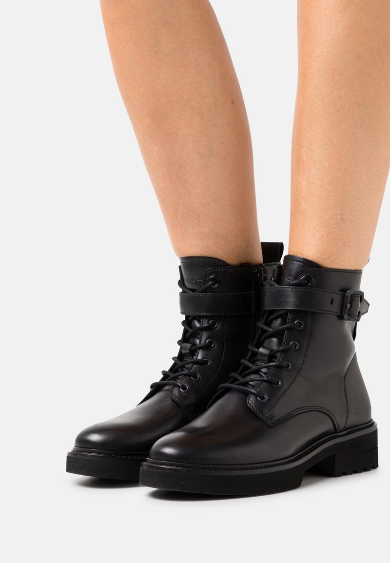 Bottes courtes en cuir noir avec un bout arrondi, lacets à l'avant, bride à la cheville et semelle en caoutchouc texturée. Présentent des accents matériels minimalistes.