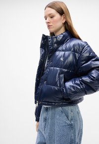Tumesinine puhv coat läikiva tekstuuriga, nööpide ja tõmblukuga kinnitused, küljetaskud ning tikandiga logo rinnal.