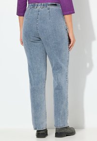 Henkilö pukeutuneena korkeavyötäröisiin vaaleansinisiin farkkuihin, violettiin toppiin ja mustiin saappaisiin, seisoo valkoista seinää vasten, kädet rentoina sivuilla.