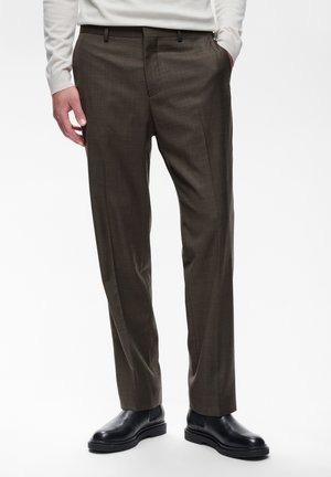 Homme portant un pantalon marron foncé sur mesure avec poches latérales et bottes en cuir noir, debout sur un fond uni.