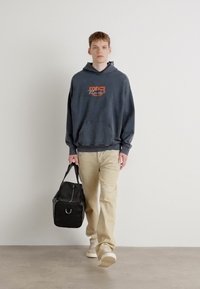 Sudadera navy con logo, pantalones beige, zapatillas blancas y bolsa de deporte negra. El conjunto presenta un corte relajado y texturas suaves.