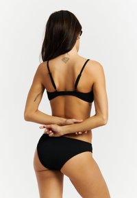 Mujer con cabello largo y oscuro, con bikini negro texturizado, brazos cruzados detrás de la espalda, mostrando un tatuaje en la parte superior de la espalda con forma de diamante.