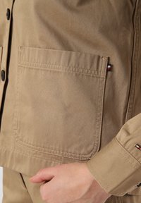 Beżowa bawełniana kurtka z kieszenią na piersi, czarnymi guzikami i małym akcentem logo. Materiał ma gładką fakturę i solidną konstrukcję.
