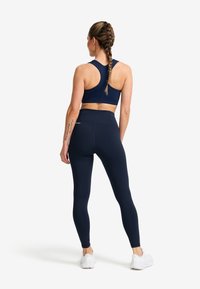Kvinna med flätat hår som bär en marinblå sportbh och leggings, stående med ryggen mot kameran på vit bakgrund.
