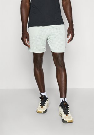 Mann trägt schwarzes T-Shirt, hellgraue perforierte Shorts, weiße Socken und weiße Sneakers mit schwarzen Schnürsenkeln und steht auf grauem Boden.