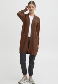 Bruine gebreide cardigan met lange mouwen, open voorkant en twee voork pockets, gecombineerd met donkere broeken en beige sneakers.