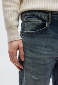 Világoskék farmernadrág szakadt részletekkel, karcsúsított szabással és kontrasztos varrással. A kéz a zsebben pihen, a viselet krémszínű, bordázott pulóverből áll.