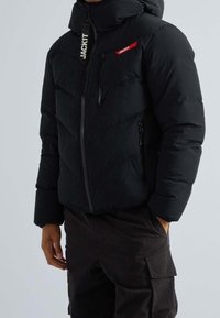 Doudoune noire avec col haut, motif matelassé diagonal, poches zippées et détails rouges avec le mot "JACKIT".