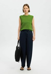 Grünes ärmelloses Strickoberteil, navy breite Hose und schwarze Netztragetasche. Das Model trägt schwarze Flip-Flops. Einfaches, lässiges Outfit.