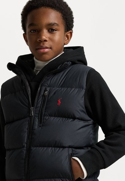 Črna puffer brezrokavnik z rdečim logotipom, z zadrgo in kapuco. Obučen čez bel polo pulover z teksturiranim, quilted dizajnom.