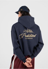 Sudadera de color navy con el diseño de "Prohibido" bordado en dorado y una rama de olivo en la parte posterior, combinada con una gorra navy a juego.