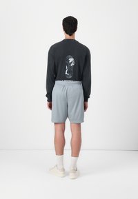 Zwart shirt met lange mouwen met een grafisch ontwerp van een schoenafdruk; gecombineerd met lichtblauwe shorts; model draagt beige schoenen en witte sokken.