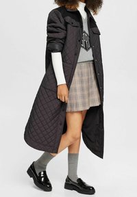 Abrigo largo negro acolchado con botones a presión, con un cuello sólido y mangas enrolladas, llevado sobre un suéter gris y una falda de cuadros. Zapatos negros y calcetines grises.