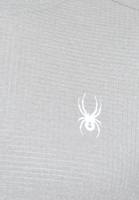 Lichtgrijs, textuurshirt met een wit spinnenlogo op de voorkant. De stof heeft een ribbelpatroon en een ronde hals.