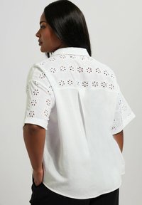 Camicia bianca a maniche corte, con ricami floreali intagliati lungo le spalle e la schiena, dal taglio morbido e con colletto.