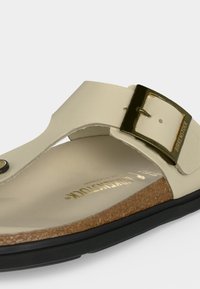Μπεζ σανδάλι Birkenstock με χρυσή αγκράφα, φελλό πάτο και μαύρη σόλα, εμφανιζόμενο σε κοντινή λήψη από γωνιακή πλευρική όψη.